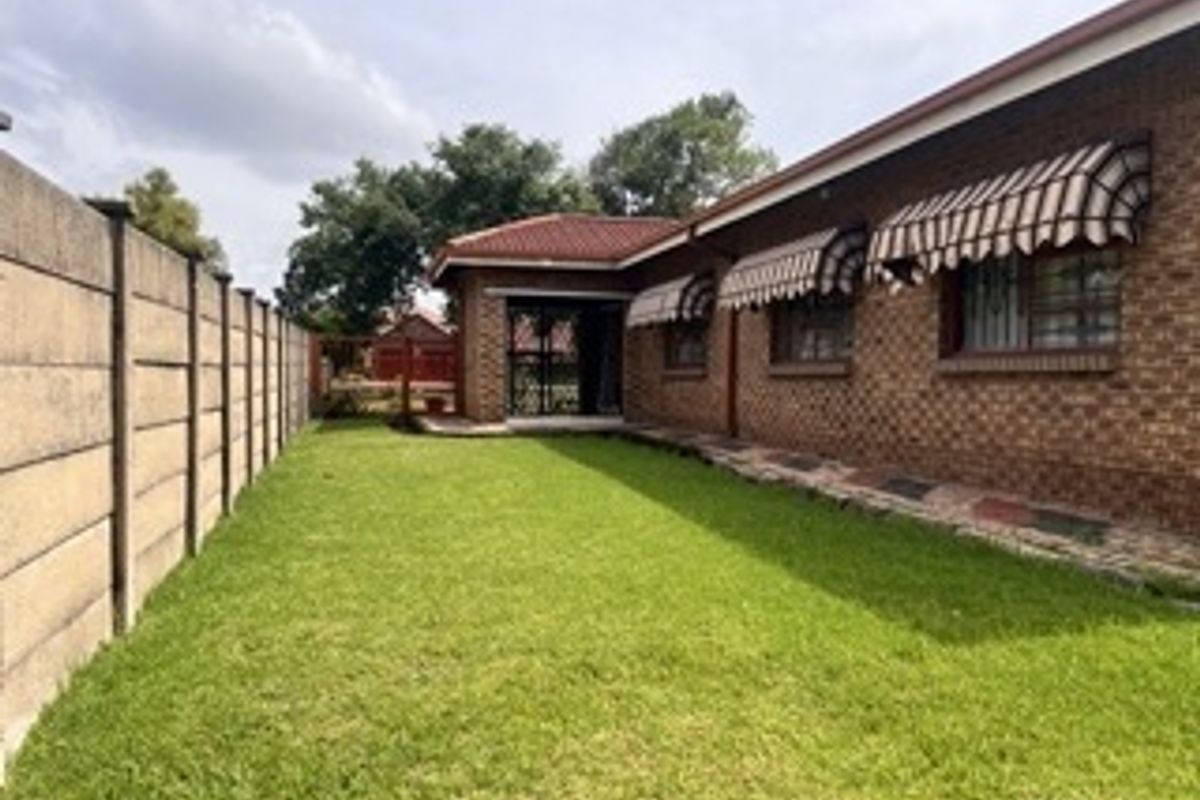 Rear view of the property featuring a brick facade, tiled roof, window awnings, and a well-maintained lawn enclosed by a precast concrete boundary wall.