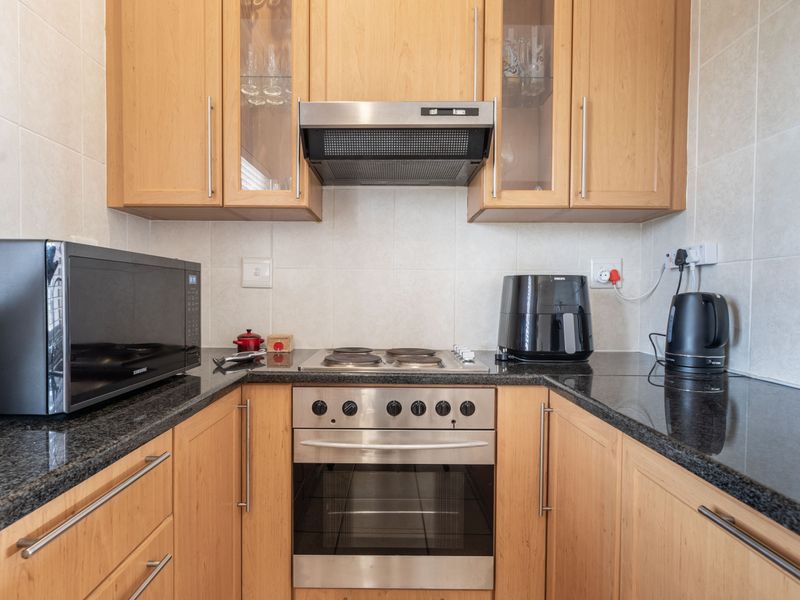 A compact, functional kitchen featuring light wood cabinetry, granite countertops, a stainless steel oven, and a built-in extractor fan with tiled backsplash.