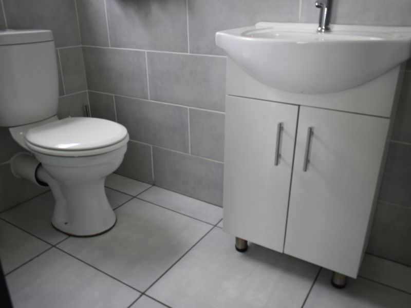 A clean, functional bathroom featuring a white toilet, a modern vanity cabinet with an integrated washbasin, and neutral grey wall and floor tiling.