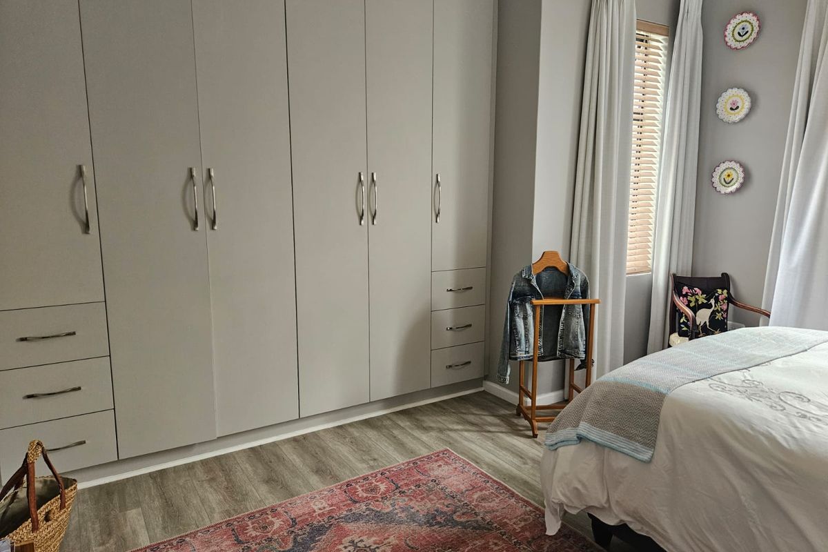A well-maintained bedroom featuring extensive floor-to-ceiling built-in cupboards, modern laminate flooring, and natural light through venetian blinds.
