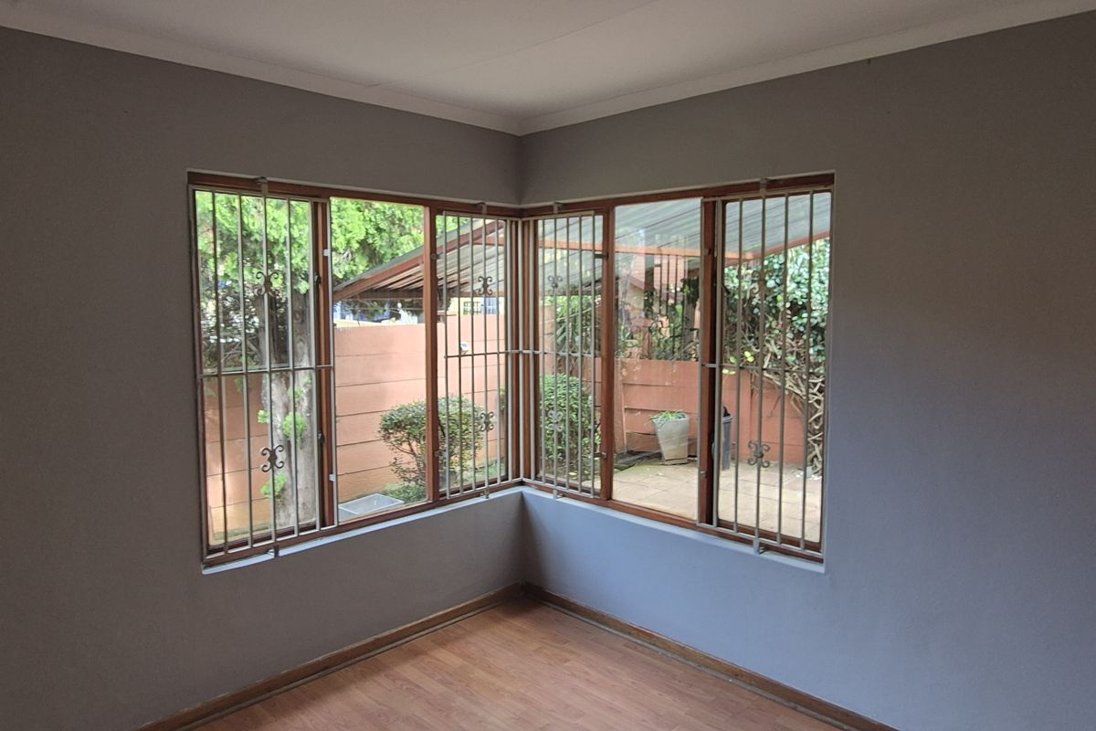 A bright, empty bedroom featuring wood-look laminate flooring, corner windows with security bars, and neutral grey walls.