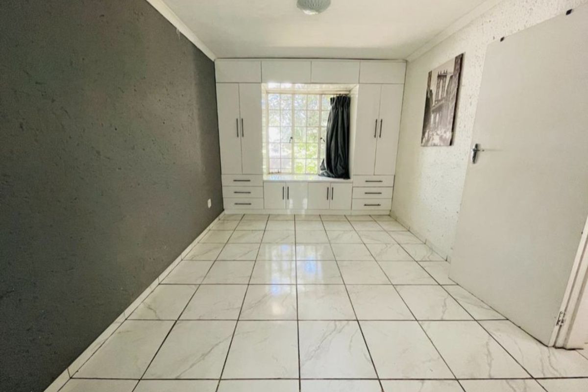 A bright, empty bedroom featuring white tiled flooring, extensive built-in cabinetry with an integrated window seat, and a contrasting dark accent wall.
