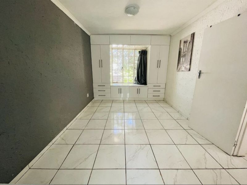 A bright, empty bedroom featuring white tiled flooring, extensive built-in cabinetry with an integrated window seat, and a contrasting dark accent wall.