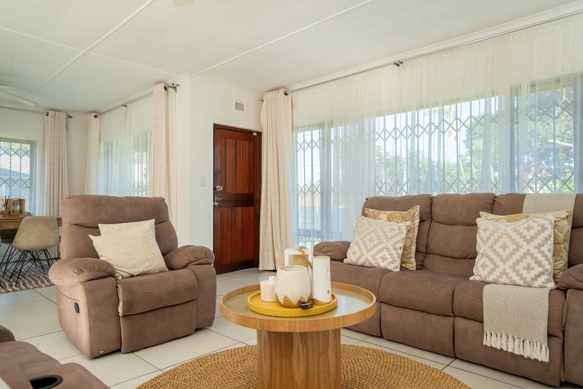 A bright and spacious living area featuring neutral tiled flooring, large windows with security gates, and direct access to the home entrance.