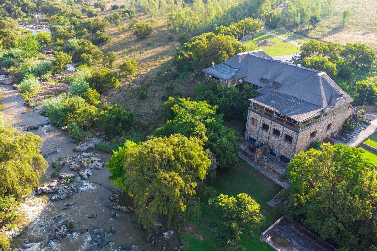 Aerial view of a multi-level stone guest house with a thatched roof, situated along a scenic riverbank surrounded by lush greenery.