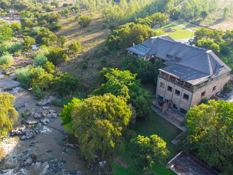 Aerial view of a multi-level stone guest house with a thatched roof, situated along a scenic riverbank surrounded by lush greenery.