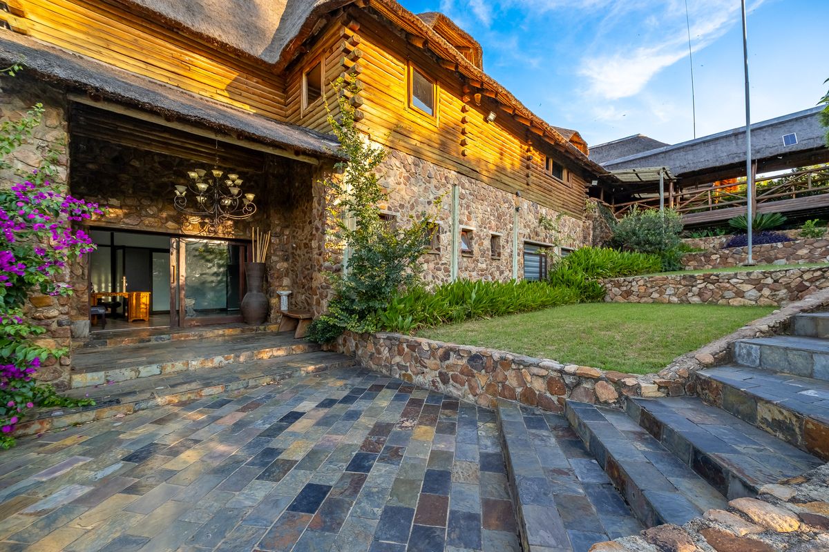 A rustic guest house exterior featuring stone and log construction, a thatched roof, slate patio, and landscaped terraced gardens with stone retaining walls.