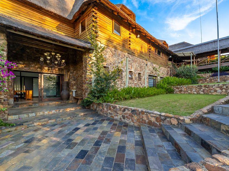 A rustic guest house exterior featuring stone and log construction, a thatched roof, slate patio, and landscaped terraced gardens with stone retaining walls.