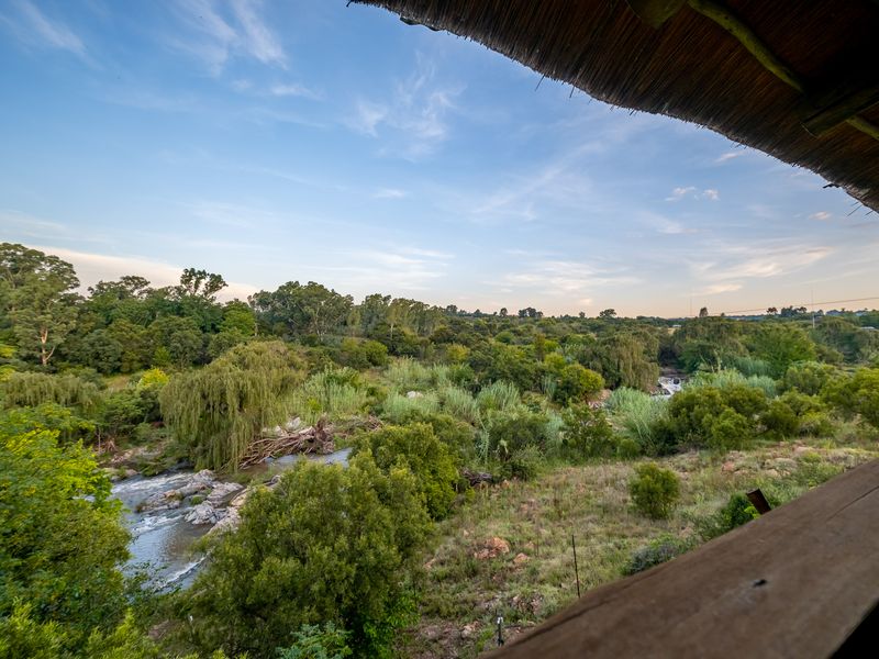 Scenic elevated view overlooking a lush river valley with dense vegetation and natural landscape, framed by a traditional thatched roof structure.
