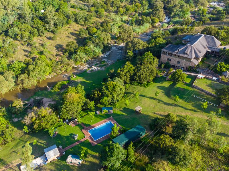 Aerial view of a guest house property featuring a swimming pool, extensive landscaped gardens, and river frontage with stone-built structures and thatched roofing.