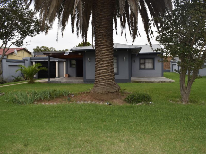 Front view of a single-story house featuring a covered patio, grey exterior finish, and a large front garden with mature palm tree.