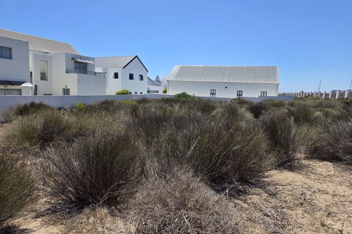 A vacant residential plot featuring sandy terrain and native shrubbery, bordered by a perimeter wall with neighboring residential properties in the background.