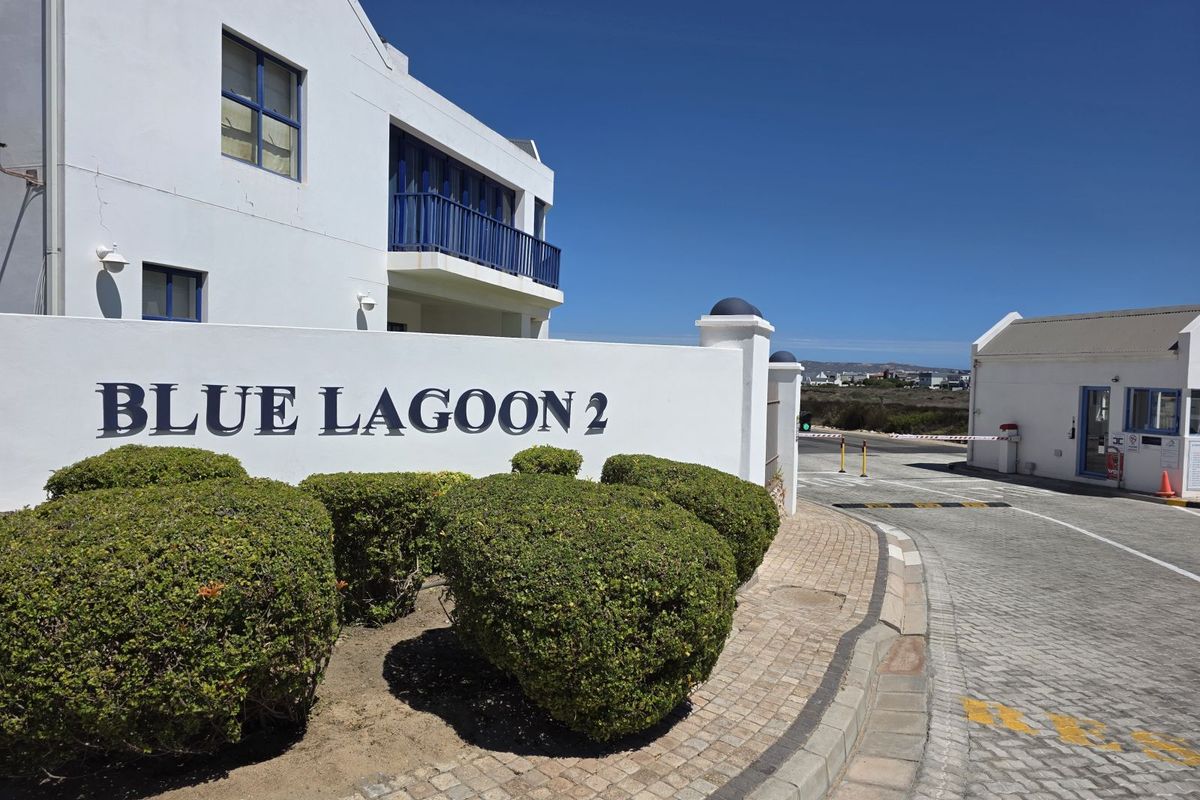 Entrance view of Blue Lagoon 2 residential estate featuring a paved access road, security gatehouse, and landscaped garden beds.