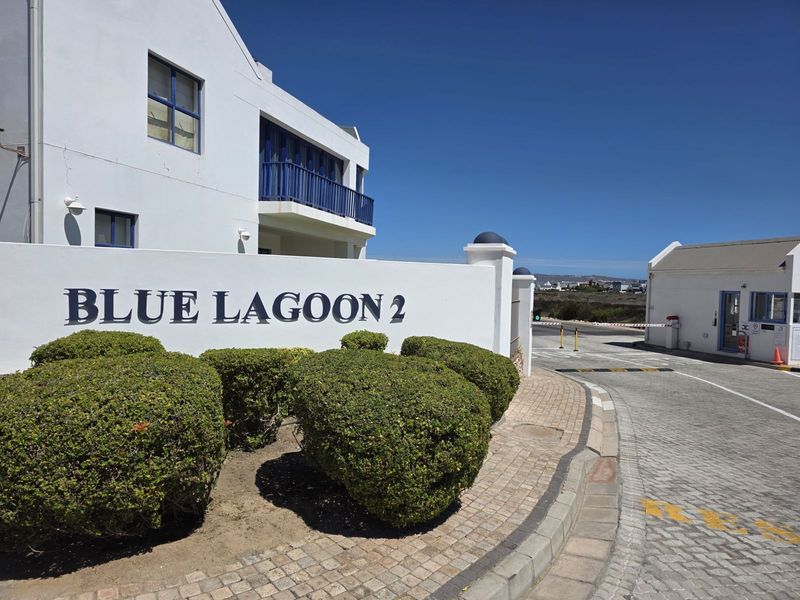 Entrance view of Blue Lagoon 2 residential estate featuring a paved access road, security gatehouse, and landscaped garden beds.