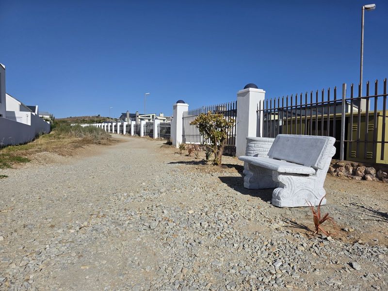 A gravel pathway bordering a residential estate, featuring a stone bench, metal perimeter fencing, and concrete pillars.