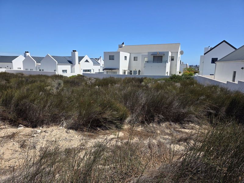 A vacant residential plot covered in natural coastal vegetation, situated within a developed residential estate with surrounding white-walled homes.
