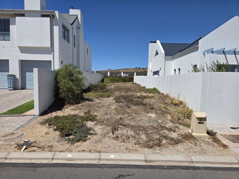 A vacant residential plot situated between two existing modern white-walled properties, featuring level ground and established boundary walls.