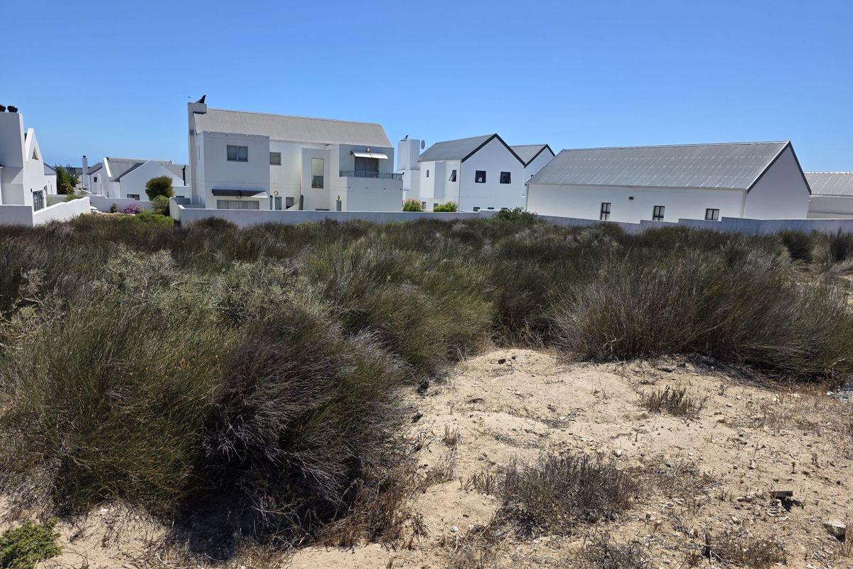 A vacant residential plot featuring natural coastal vegetation and sandy terrain, situated in a developed neighborhood with established white-walled homes.