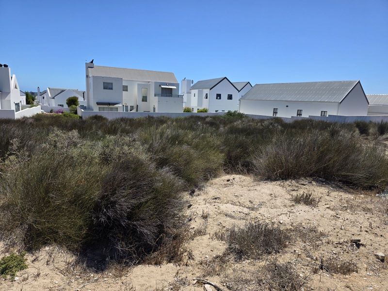 A vacant residential plot featuring natural coastal vegetation and sandy terrain, situated in a developed neighborhood with established white-walled homes.