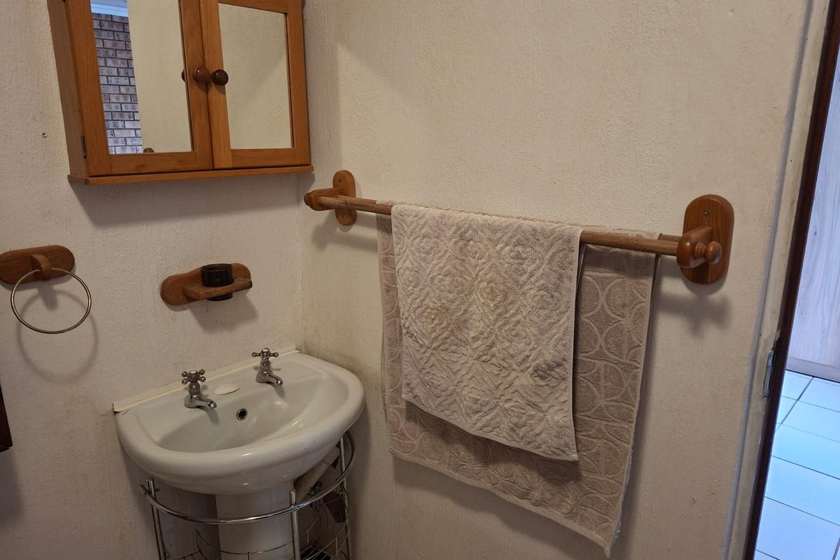 A compact bathroom featuring a pedestal basin, wooden medicine cabinet, and matching wooden towel rail, showcasing dated finishes in need of modernization.