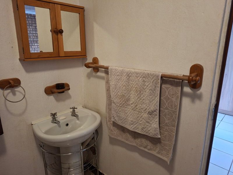 A compact bathroom featuring a pedestal basin, wooden medicine cabinet, and matching wooden towel rail, showcasing dated finishes in need of modernization.