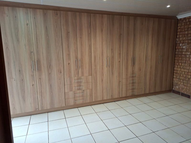 A spacious bedroom featuring extensive floor-to-ceiling built-in wooden cabinetry, ceramic tiled flooring, and a decorative exposed brick feature wall.