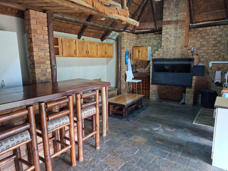 Rustic outdoor entertainment area featuring a thatched roof, exposed brick walls, stone flooring, and a built-in braai, ideal for social gatherings.
