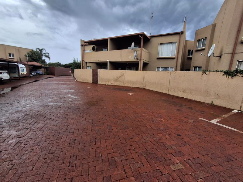 Exterior view of the townhouse complex featuring a paved driveway, boundary wall, and covered parking area under an overcast sky.