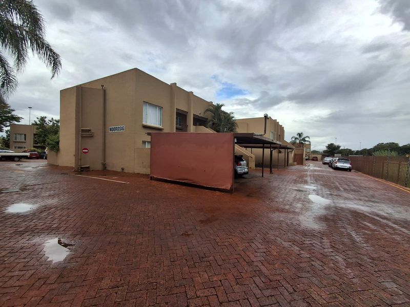 Exterior view of the townhouse complex featuring a paved driveway, covered carport parking, and perimeter fencing under an overcast sky.