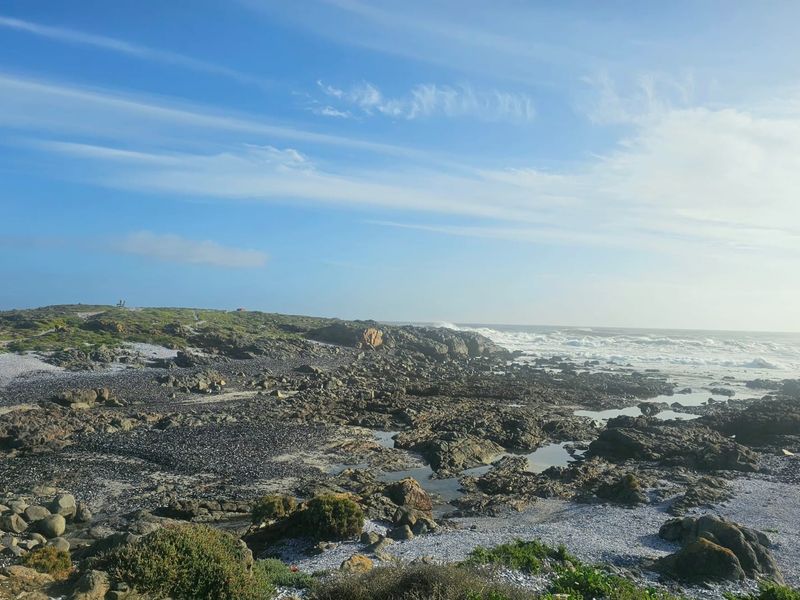 A scenic coastal view featuring rugged rocky terrain, natural shoreline, and ocean waves under a clear blue sky.