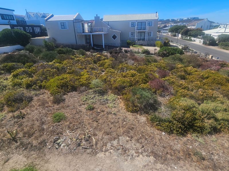 An elevated view of a vacant residential plot featuring natural coastal vegetation, situated in a developed neighborhood with nearby street access.