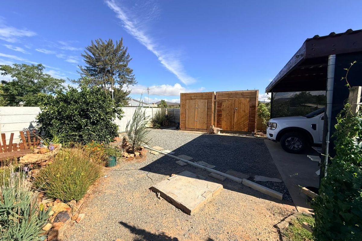 A rear outdoor area featuring a gravel driveway, wooden storage units, and a covered parking space adjacent to the main dwelling.