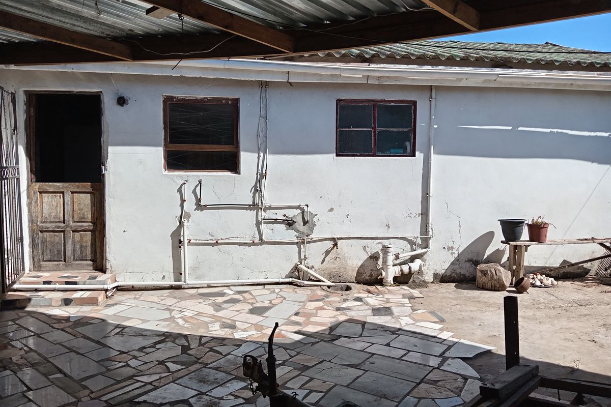 Rear exterior area featuring a covered patio with crazy paving, exposed plumbing, and weathered wall finishes requiring maintenance.