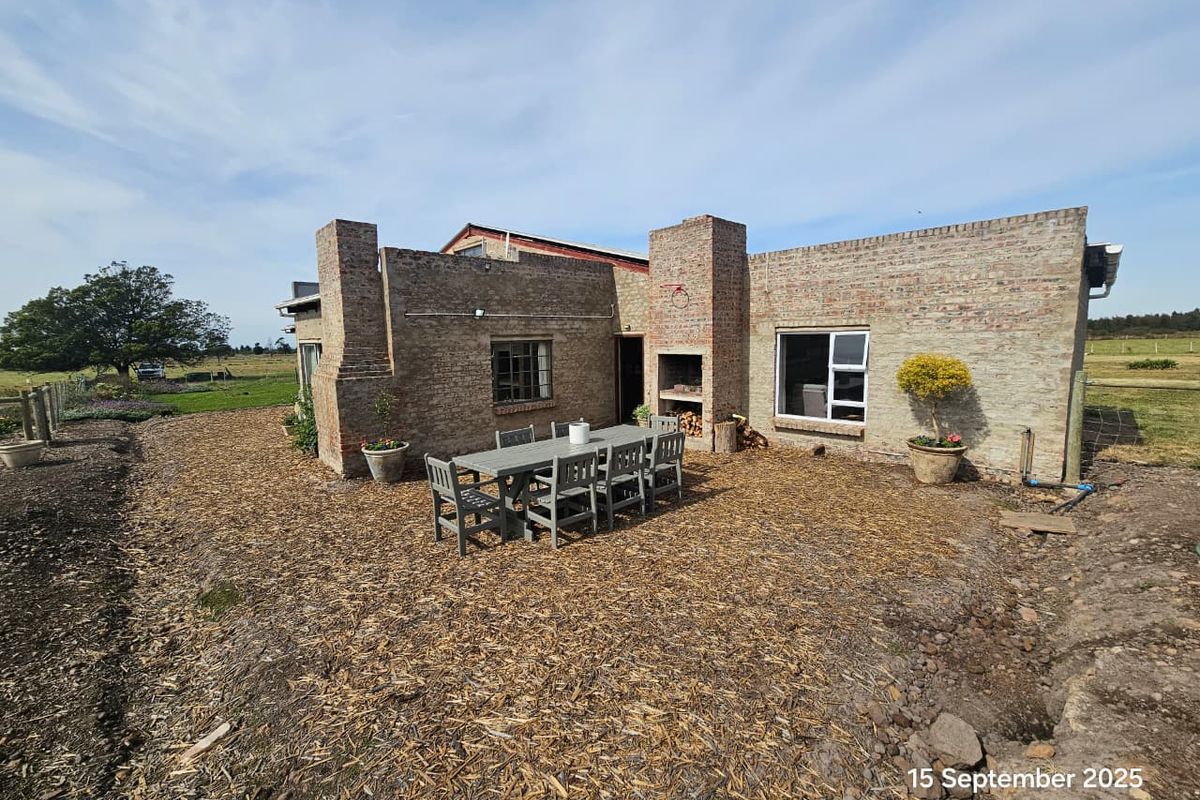 Rear exterior view of a rustic brick farmhouse featuring a built-in outdoor fireplace and a spacious patio area overlooking the surrounding farmland.