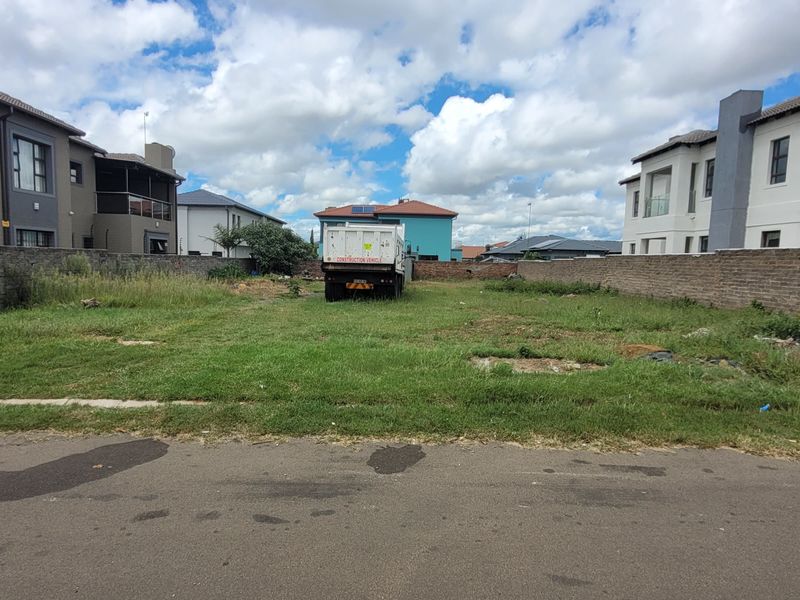 A vacant residential plot situated between existing modern homes, featuring a level grass surface and perimeter boundary walls.