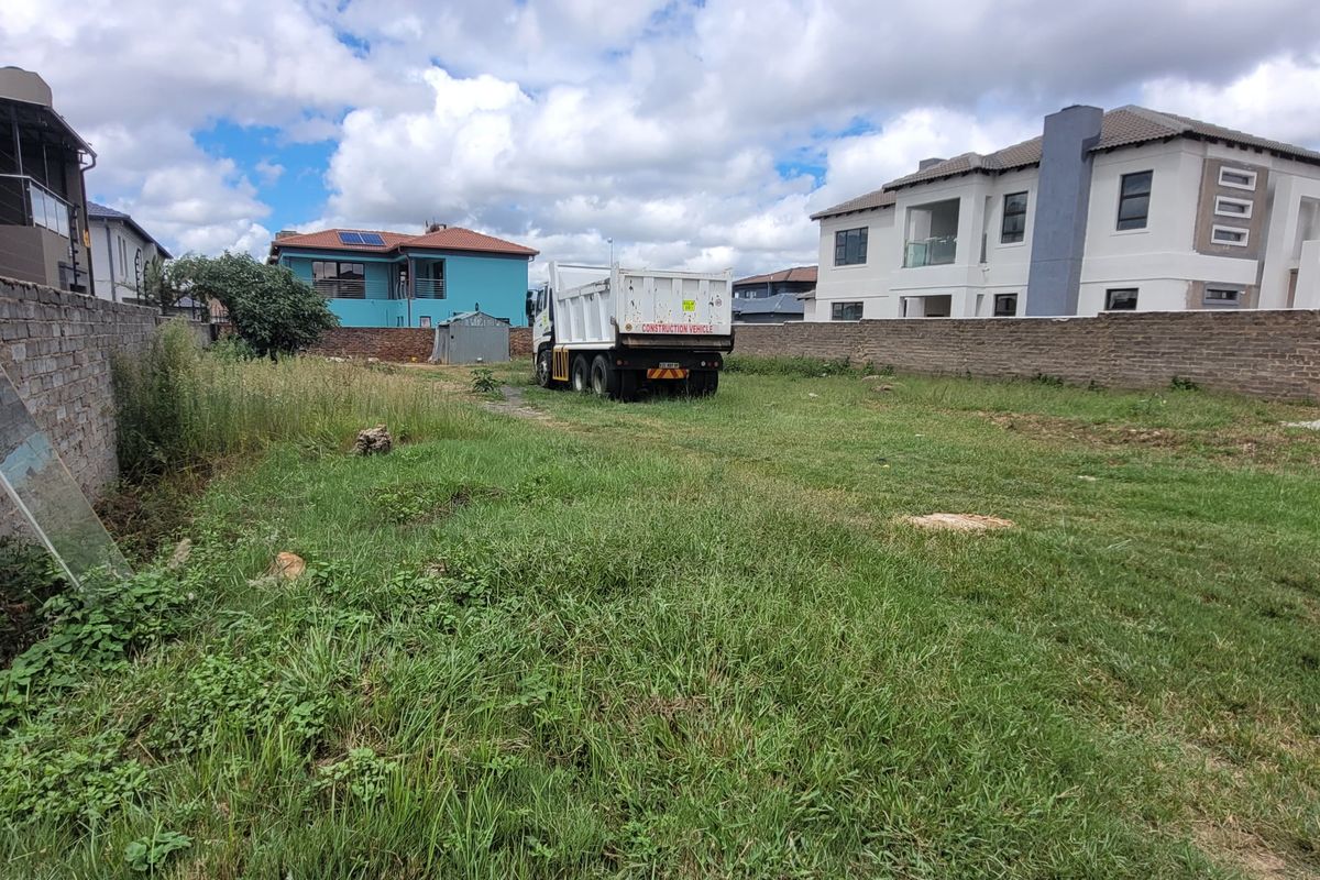 A vacant residential plot enclosed by perimeter brick walls and electric fencing, featuring overgrown grass and surrounding residential developments.