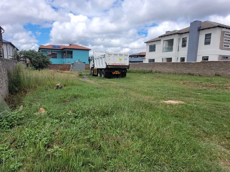 A vacant residential plot enclosed by perimeter brick walls and electric fencing, featuring overgrown grass and surrounding residential developments.