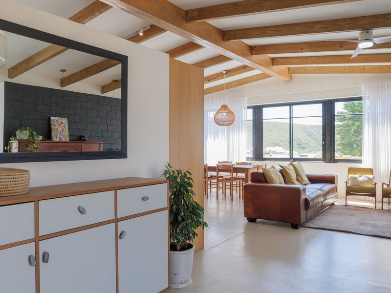 Bright open-plan living and dining area featuring polished concrete floors, exposed timber ceiling beams, and a striking black brick feature wall with large windows.