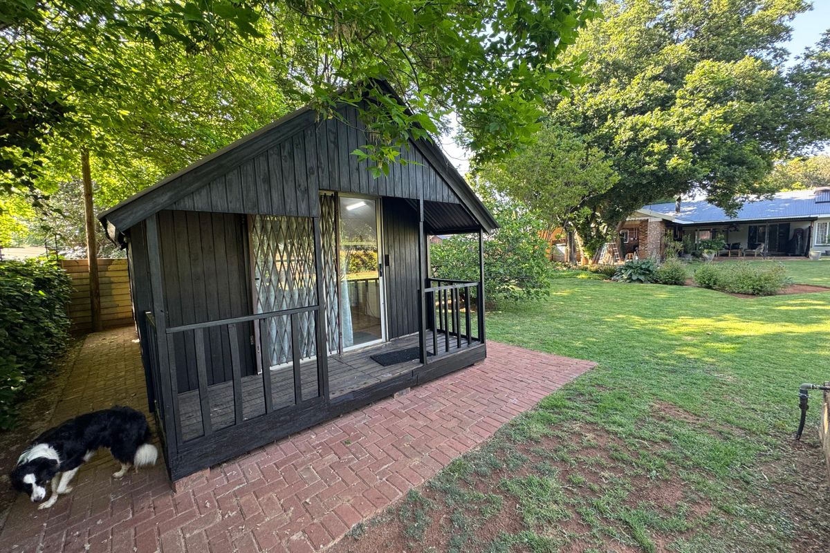 A detached timber cabin featuring a covered porch and sliding door access, situated in a well-maintained garden setting with brick paved pathways.