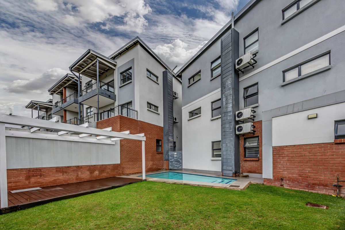 Rear view of the apartment complex featuring a communal swimming pool, wooden deck, pergola, and private balconies with exterior air conditioning units.