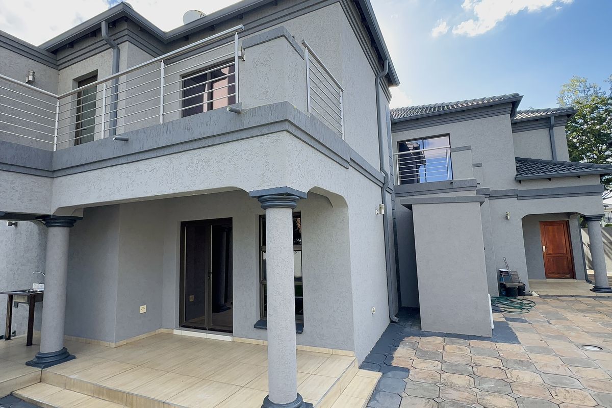 Rear view of a modern double-story house featuring a covered patio, upper-level balcony with stainless steel railings, and a paved courtyard area.