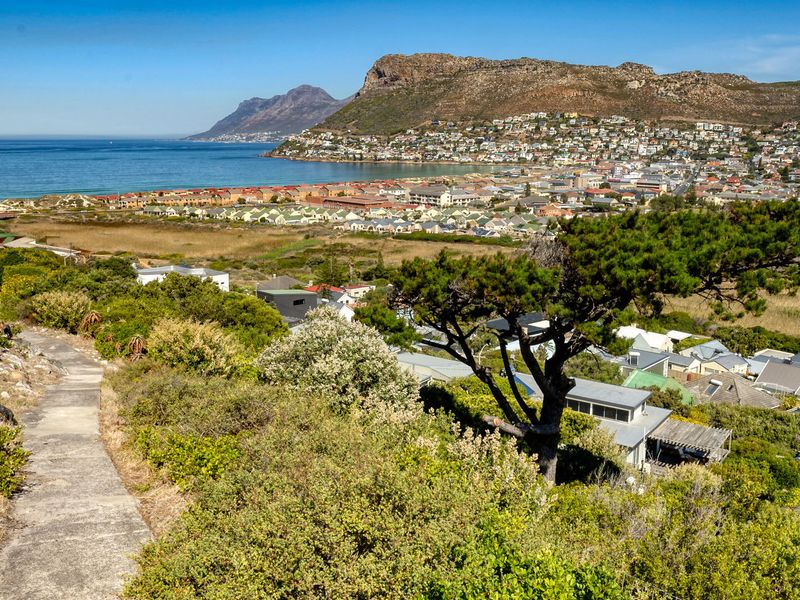 Elevated panoramic view overlooking the coastal town, bay, and surrounding mountain landscape, featuring a concrete pathway leading through the natural hillside vegetation.