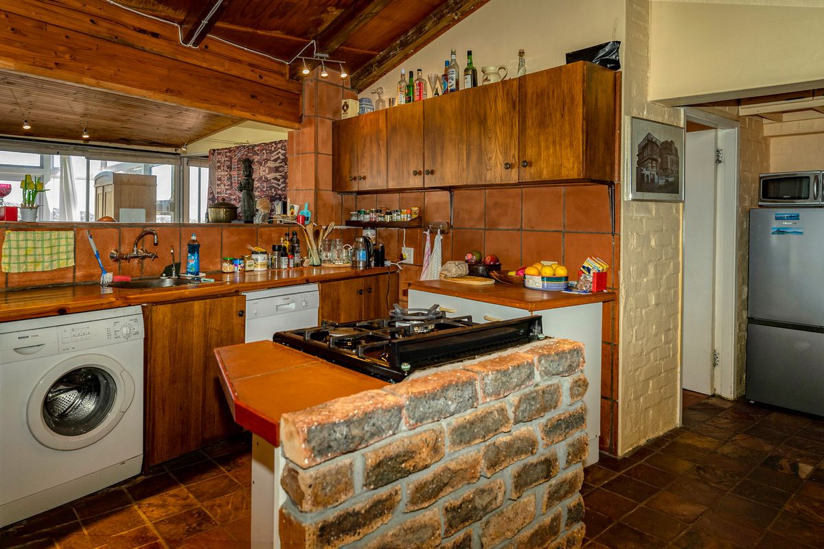 A rustic kitchen featuring wooden cabinetry, a brick-based island with a gas hob, terracotta tile backsplash, and exposed timber ceiling beams.
