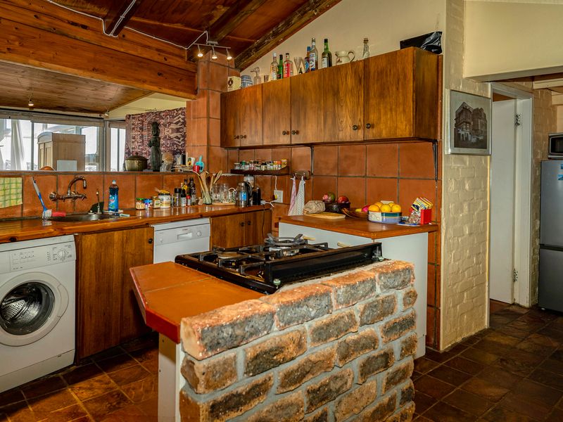 A rustic kitchen featuring wooden cabinetry, a brick-based island with a gas hob, terracotta tile backsplash, and exposed timber ceiling beams.