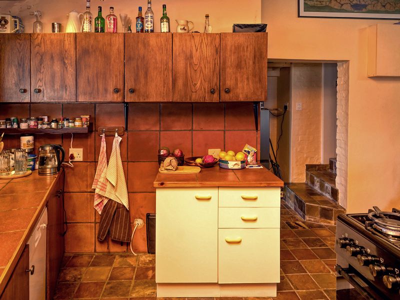 A rustic kitchen featuring wooden cabinetry, tiled surfaces, and a built-in gas stove, with a stone feature wall and access to a secondary area.