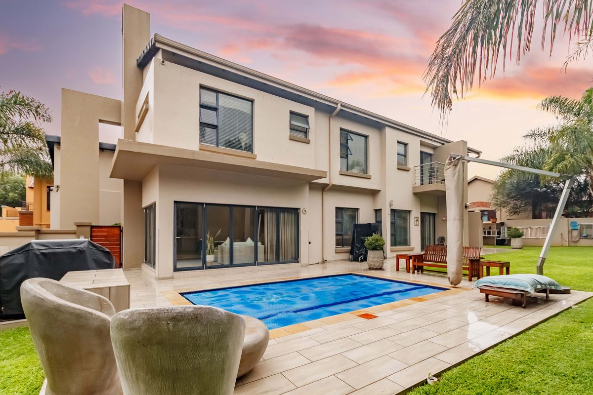 Rear view of a modern double-story house featuring a swimming pool, tiled patio area, and balcony, set within a landscaped garden.