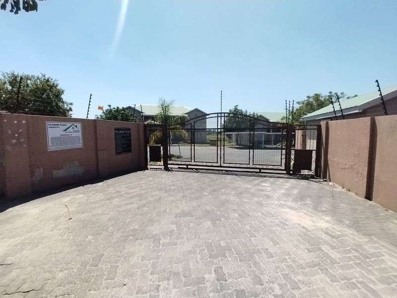 Front entrance view of the apartment complex featuring a secure metal gate, paved driveway, and perimeter wall with electric fencing.