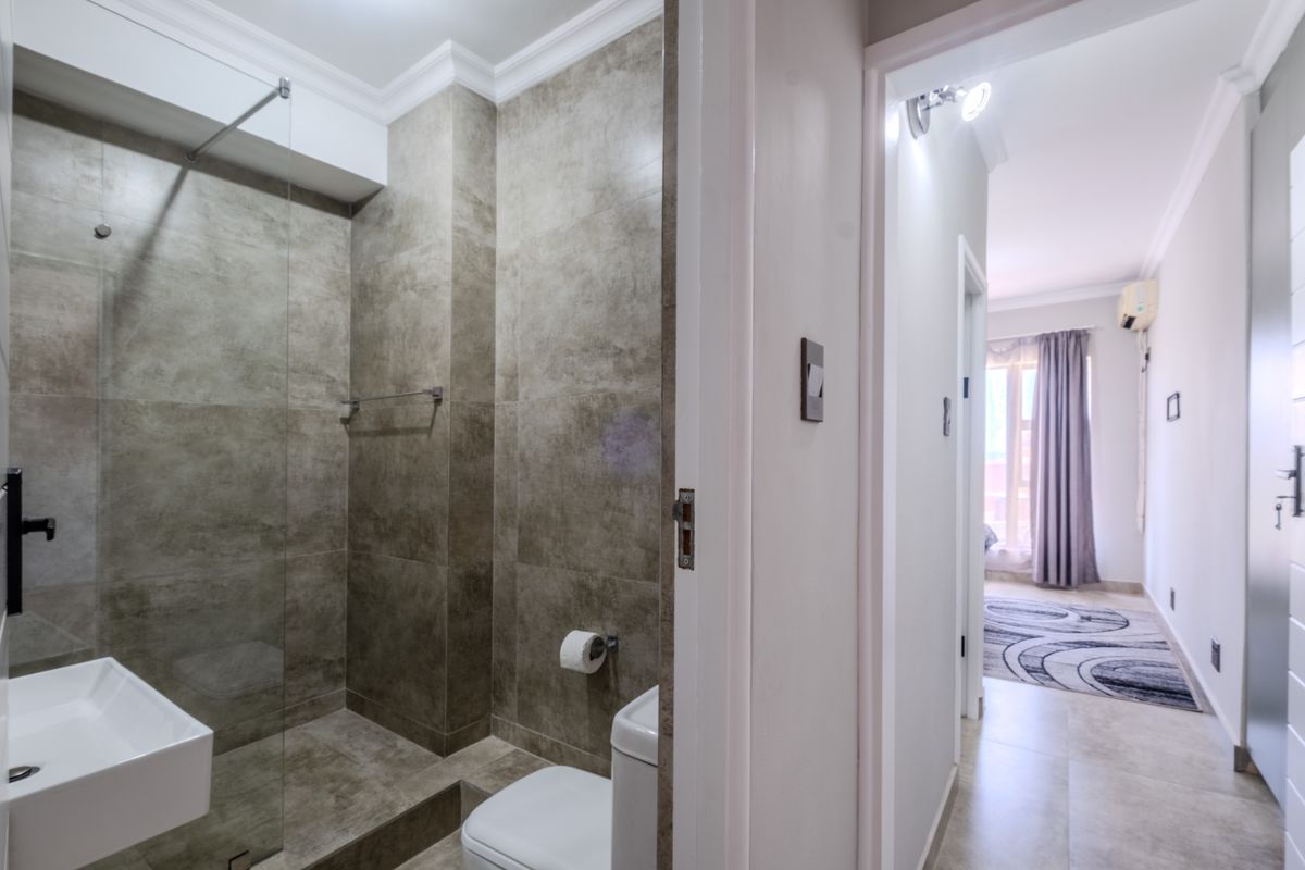 Modern bathroom featuring a glass-enclosed shower with large-format grey tiles, adjacent to a hallway leading into a bright bedroom.