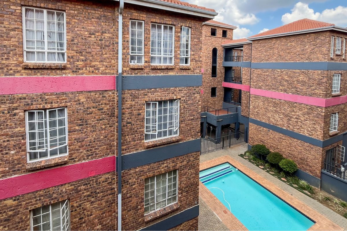 An elevated view of an apartment complex courtyard featuring a swimming pool, paved deck, and multi-story brick buildings with painted accents.