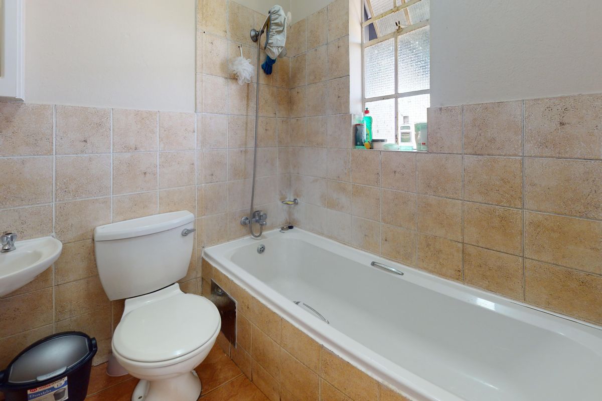 A dated bathroom featuring a pedestal basin, toilet, and bathtub with wall-mounted shower mixer, finished with neutral-toned wall and floor tiles.